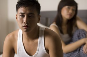 An asian man looks unhappy sitting on the bed, with his partner (out of focus) behind him